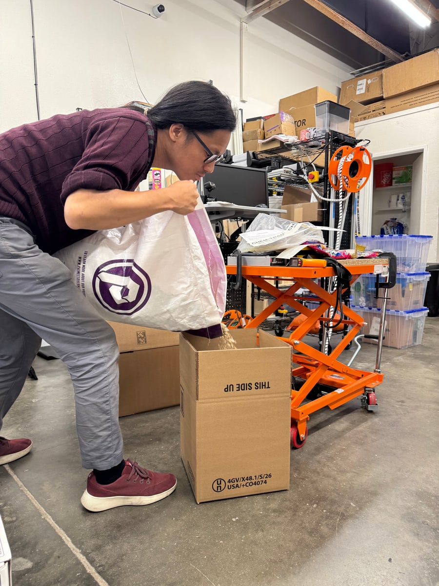 A person pours granules from a large bag into a cardboard box beside an orange lift table.