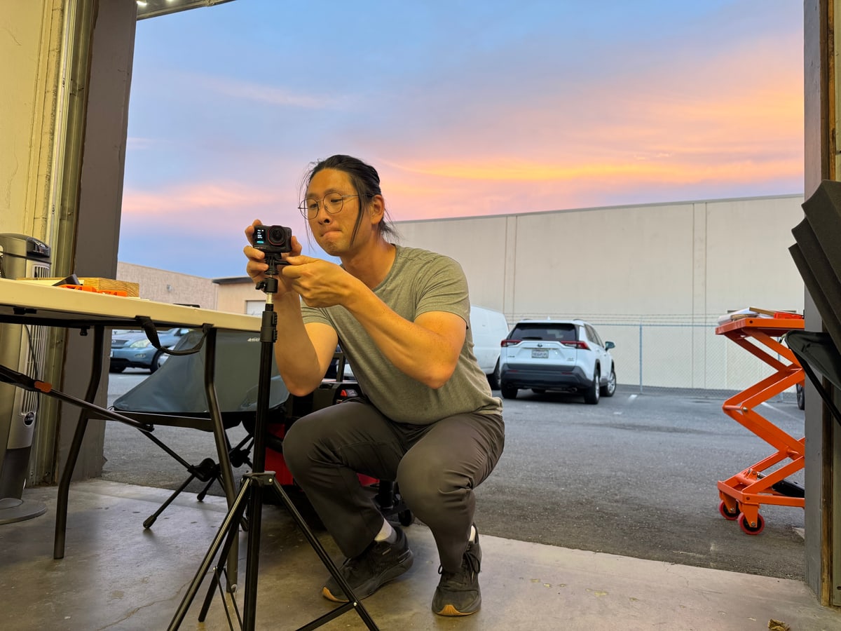 A person adjusts a camera on a tripod at an open workshop door at sunset.