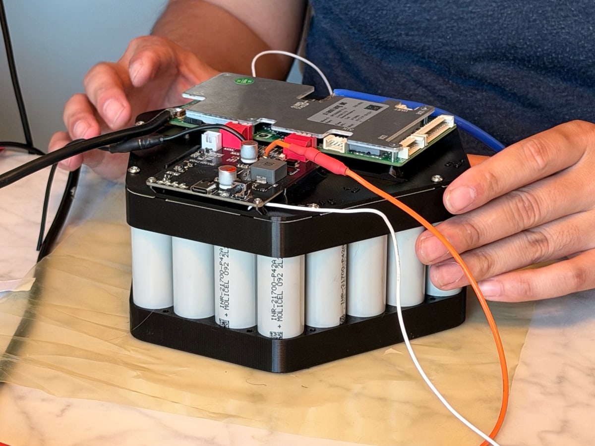 Hands work on circuit boards mounted above a pack of cylindrical battery cells.